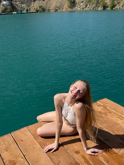 A young woman with long blonde hair, wearing a white crocheted bikini, is lying on a wooden dock. Her eyes are closed, and she is smiling broadly, conveying a sense of joy and relaxation. Her hair is spread out behind her, and her left arm is extended, resting on the dock. The dock is made of weathered wooden planks and is positioned at the edge of a body of bright turquoise water. The water appears calm with subtle ripples. In the background, to the left of the water, is a steep, rocky embankment with sparse vegetation. Further down the embankment, a small structure with white stairs leading to the water can be seen. The lighting suggests it is a sunny day, likely late morning or early afternoon, as indicated by the strong shadows cast on the dock. The overall scene is idyllic and evokes a feeling of summer leisure.