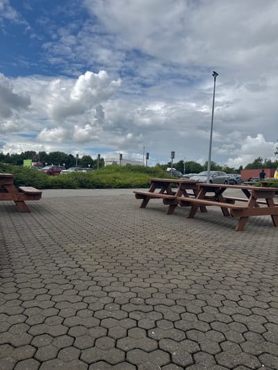 This is a rotated, landscape photograph of an outdoor seating area with picnic tables in a paved parking lot. The paving stones are hexagonal and dark gray. Two wooden picnic tables with attached benches are positioned in the foreground, angled towards the viewer. They are made of dark brown wood. Beyond the tables, several cars are parked in a lot, bordered by a dense line of green bushes and trees. In the background, a large shipping container, possibly red, is visible.

The sky is filled with dramatic clouds. On the left, a large, fluffy white cloud dominates the frame against a bright blue sky. On the right, darker gray clouds are present, suggesting a mix of sun and possible rain. A tall, slender lamppost stands on the left side of the image, extending into the sky.

The image appears to have been taken during the daytime. The weather seems to be partly cloudy. The scene is likely a rest stop, a public park, or a service area with outdoor amenities. There is no discernible activity taking place in the image beyond the presence of the parked cars. No people are clearly visible or engaged in any action. The overall mood is somewhat neutral, capturing an ordinary outdoor scene. No visible text is present in the image. The location context is Commons Cross, Ireland.