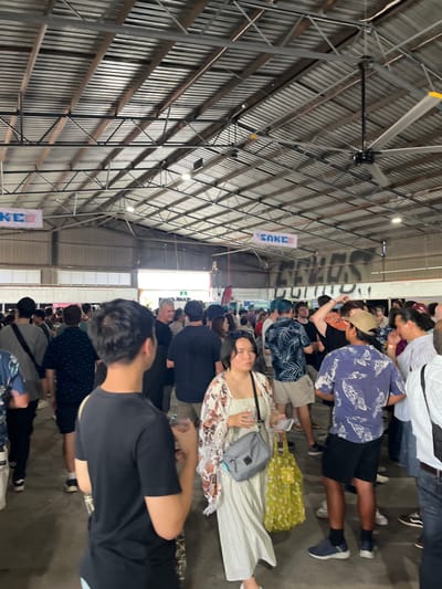 The image shows a large crowd of people gathered inside a large, metal-roofed building. The roof is supported by a network of metal beams and there are several large ceiling fans. The building is lit by fluorescent lights and has a concrete floor. The crowd is mostly standing and some people are holding drinks. The image appears to have been taken at the Hamilton Food and Wine Festival, held annually at the Hamilton Showground in Hamilton, Queensland, Australia (latitude: -27.44, longitude: 153.073). A banner reading "SAKE" is visible in the background.  The scene is bustling with activity and a sense of community. The people are dressed casually in everyday attire and there is a mix of ages and ethnicities present. The image captures a snapshot of a social event where people are enjoying themselves and connecting with each other.