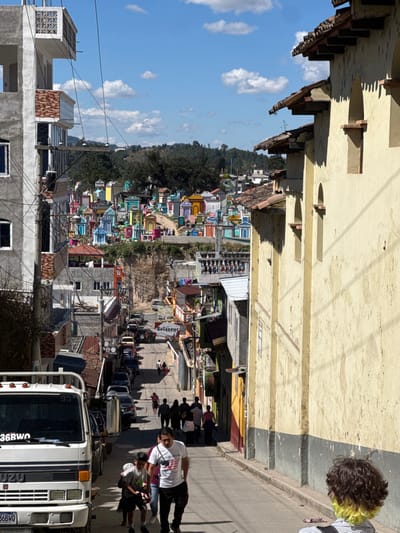 A daytime street scene, viewed at an unusual angle, features a road lined with a colorful cemetery atop a hill. Buildings border the road, with people walking by. A white truck parked on the street partially obscures the view. Blue sky with white clouds is visible above the scene, while a person with yellow-tipped hair stands on the street.