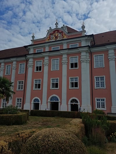 The image depicts the Neues Schloss, also known as the Prince-Bishop's New Palace, located in Meersburg, Germany. The building features a salmon-pink facade with white architectural details, including columns and window frames. The upper part of the building displays an ornate pediment with a coat of arms and decorative statues. Numerous windows are visible on different floors, some open and some closed. In the foreground, a meticulously landscaped garden with neatly trimmed hedges and various plants is present. A palm tree is visible on the left side of the image. A man in a plaid shirt is seen standing near an open doorway on the ground floor. The sky above is partly cloudy.