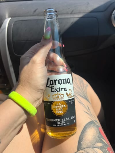 A hand with gradient pink and green fingernails holds a glass bottle of Corona Extra beer. The hand is adorned with a bright yellow wristband. The bottle, partially filled with golden liquid, is labeled with the Corona Extra logo. The background appears to be the interior of a vehicle, with a dark dashboard and a sunlit area. The person's tattooed leg is visible in the lower right portion of the image. The location is identified as Novye Shal'tyamy, Russia.