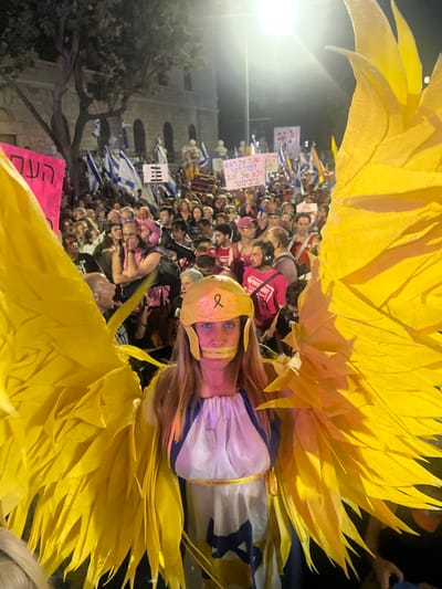 An adult female stands prominently in the foreground, facing the viewer directly amidst a large nighttime street protest. She wears a white and blue toga-like garment featuring a blue Star of David symbol on its lower front, accented with a yellow ribbon around her waist that bears the text "7.10.2023". She is adorned with large, structured yellow feathered wings and a yellow helmet, which displays a dark ribbon symbol on its front. Yellow tape covers her mouth.

Behind her, a dense crowd of people, varied in age and gender, fills the street. Many hold up numerous Israeli national flags (blue Star of David on white). Several protest signs are visible, including one pink sign with partially visible Hebrew text "העם..." (The People...), and another white sign with a unique black symbol of three horizontal lines intersected by a vertical line. A pink sign in the background states in Hebrew: "שנה שראוי ... ולא של נכד ביביסט" (A year that is worthy... and not of a Bibi-ist grandson). The scene is illuminated by overhead streetlights. In the far background, a multi-story stone building with arched windows and classical architectural elements is visible.

This protest scene is situated on a city street in West Jerusalem, Israel.