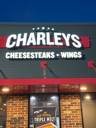 This is a vertical, eye-level, outdoor, medium shot of the Charleys Cheesesteaks and Wings restaurant facade at night, with a clear dark blue sky above. The building's exterior is dark grey with horizontal siding. Above the entrance, a large, illuminated white sign reads "CHARLEYS" in all caps, with four white stars above the letters. Below this, another illuminated white sign reads "CHEESESTEAKS • WINGS." Red trim is visible above and below the signs. The entrance area features red brick walls and large windows, with some interior lights visible. A poster inside the window promotes a "LIMITED TIME! THE OLD SCHOOL TRIPLE MELT."