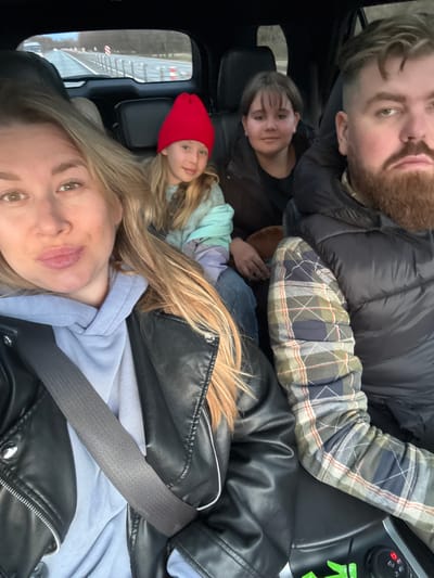 Four people are in a vehicle. From the perspective of the camera: a blonde woman in a black leather jacket and blue hooded shirt is in the front; a younger woman in a red knit cap is behind her; further back is a person with short dark hair. In the far back corner is a bearded man in a dark puffer coat and plaid shirt. The camera is likely held by the woman in the front. The location appears to be near a road, possibly a highway, in Gubarevo, Russia. There is no definitive evidence to confirm the specific location as Gubarevo, Russia.