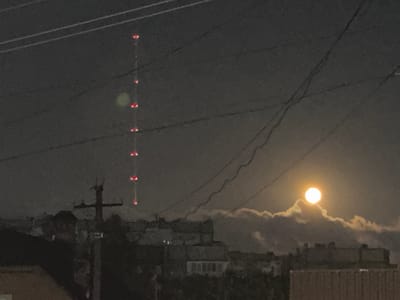 This media file depicts a nighttime scene in Obninsk, Russia, dominated by a tall radio tower with red lights and a full moon partially obscured by clouds. The setting is urban, with buildings visible in the foreground and mid-ground, some of which have illuminated windows. A utility pole with crossbars is prominent in the left mid-ground, with thick wires crisscrossing the sky. The moon, a bright, luminous orb, is positioned to the right of the frame, peeking through a bank of clouds that are lit by its glow. The overall atmosphere is somewhat hazy and atmospheric, with the dark sky punctuated by the man-made lights of the tower and the natural light of the moon. The time of day is clearly night. There is no visible text.