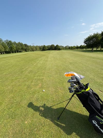 The image shows a golf bag with clubs resting on a green putting green. The bag has a black base and a yellow and black accent color, with a black strap. The golf clubs have a black and silver finish. There is a single white golf ball on the green in front of the bag. The green is in the middle of the picture, with trees on the far left and right sides. A long, dark shadow stretches to the left of the golf bag, in front of the ball. The sky is clear and blue. This scene is located in the city of Pocklington, United Kingdom, at the latitude: 53.942, longitude: -0.758.