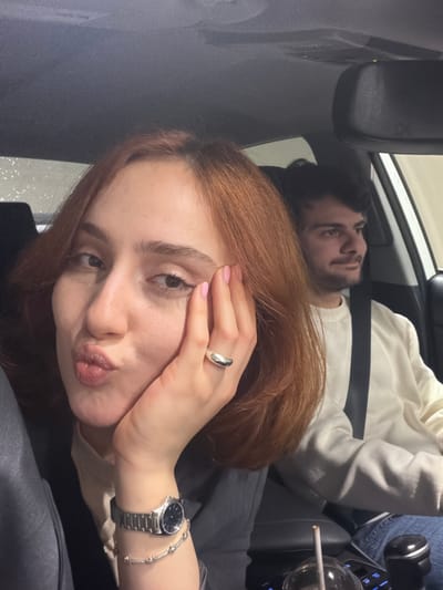 The image depicts two people inside a car. A woman with auburn hair and a silver watch and ring is in the foreground, posing with puckered lips. A man is visible in the background, lying down with a seatbelt fastened. A lit cigarette rests on the center console near a beverage container. The car's interior features a rearview mirror and dashboard components. The location is not discernible in the image.