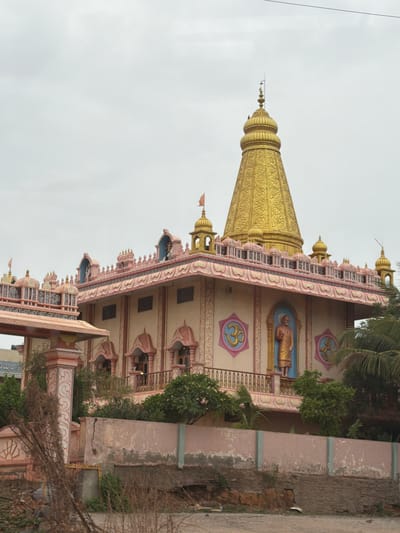A medium shot captures a temple with a gold-colored spire, located in Bethamcherla, India. The temple features pink and beige architectural elements, including ornate balconies and decorative motifs. A statue of a robed figure stands on the second story, flanked by circular emblems depicting the "Om" symbol. The building is adorned with flags and what appear to be small stupas on its roofline. In front of the temple, a low wall made of concrete and stone separates it from a dry, unpaved ground area. Vegetation, including palm trees and other foliage, is visible on the right side of the image. The sky is overcast with a pale grey hue.
