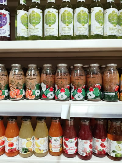 This media file features a close-up, eye-level shot of several shelves filled with various bottles of juice and flavored drinks. The bottles are arranged in neat rows, showcasing a variety of brands and flavors.
The top shelf displays bottles of "Bienky" brand carbonated flavored drinks, primarily in green apple flavor. These bottles have distinctive green labels with white text and graphics of apples.
Below, the second shelf holds bottles labeled "Smoothie." These appear to be fruit smoothies in flavors like banana-orange, apple-strawberry, and peach-apricot, as indicated by the fruit illustrations on the labels.
The bottom shelf contains bottles from "La Fruitière" brand, featuring juices like Tropical, Pomegranate (Grenade), and what appears to be apple or peach juice. These bottles have colorful labels depicting the fruits corresponding to their flavors.
The lighting suggests an indoor setting, likely a store or a pantry, with bright, even illumination. The focus is sharp on the bottles, highlighting the product details and branding. There are no people visible in the image. The scene is static, focusing solely on the display of beverages. The visible text indicates multiple languages, including Arabic, French, and English, suggesting a diverse market or multilingual packaging. There are no discernible time of day or weather cues.