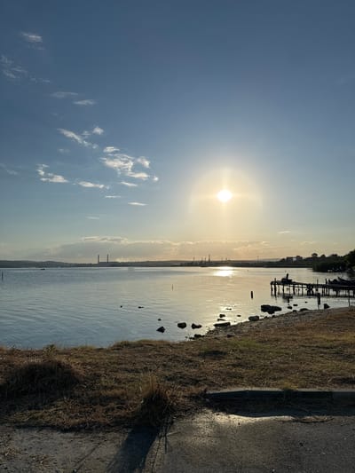 The image depicts a serene sunset scene over a body of water, likely a lake or a bay, with a distant industrial complex visible on the horizon. The sun, a bright orb, is positioned in the upper center of the frame, casting a warm, golden glow across the water and the sky. The sky is a clear blue, with a few wispy clouds scattered across it.
The foreground of the image shows a grassy, unkempt shoreline with scattered rocks. A dark, asphalt-like path runs along the bottom edge of the frame. A small, wooden pier extends into the water from the right side of the image, with a chair or bench placed on it. In the distance, the industrial complex features several tall, smokestack-like structures, suggesting a power plant or factory. The water is calm, with gentle ripples reflecting the sunlight. There are no people visible in the image.
The scene is likely located in the vicinity of Казашко, Bulgaria, an area known for its industrial activity and proximity to water bodies like Lake Varna and the Black Sea. The presence of industrial structures on the horizon strongly suggests this geographical context. The overall atmosphere is one of tranquility and industrial grandeur.