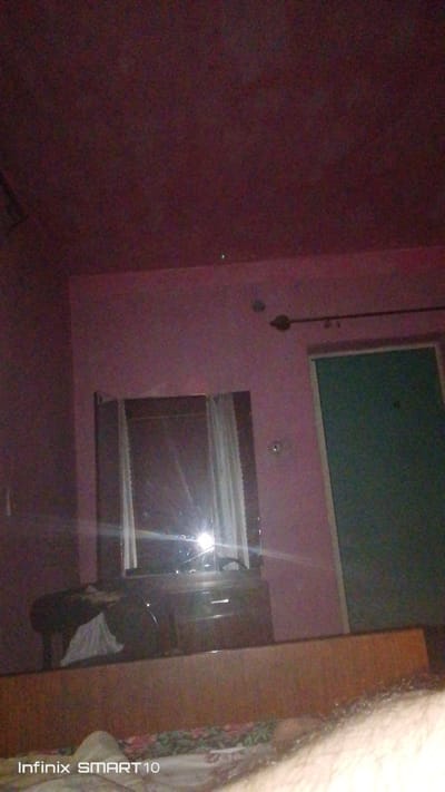 The image depicts a dimly lit room. A dresser with a mirror is visible, reflecting a window with white curtains. To the right of the mirror, a door with a dark green panel is visible. The walls appear to be painted a pinkish hue, and the ceiling is a darker reddish-purple. In the foreground, a portion of a bed with patterned bedding is visible. A watermark at the bottom left corner reads "Infinix SMART 10". The scene is consistent with an interior residential setting, potentially a bedroom, in Dustabad (Soldatsky), Uzbekistan.