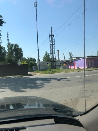 The image shows a view from inside a vehicle, looking out at a street scene with industrial and commercial buildings under a clear blue sky. Two tall, lattice cell towers are visible in the background, one on the left and a taller one on the right. Between them, a shorter industrial chimney with a metal structure around its base is also present. To the right of the chimney, a low-rise building with a predominantly purple facade and pink trim is visible, with red doors. Further to the right, a light-colored brick building with a gray roof is seen. In the foreground, the hood and windshield of the vehicle are visible, with reflections of trees and the sky on the windshield. A dark brown fence runs along the left side of the road. The road itself appears to be asphalt with white lane markings and some potholes. The overall environment suggests an urban or semi-industrial area. This scene is located in Maksimovo, Russia.