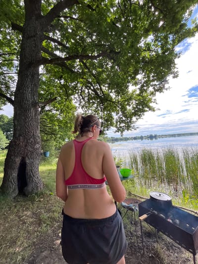 In an outdoor scene, a woman is positioned in the foreground, facing away from the viewer and towards a body of water. She is wearing a pink sports bra and black shorts. Her back is visible, and her hair is pulled back. To her right, a metal barbecue grill is set up, with smoke rising from it, suggesting food is being cooked. A large green bucket is visible to the left of the grill.
Behind the woman, a calm lake or large river stretches into the distance, reflecting the sky and surrounding vegetation. The far bank of the water is lined with tall reeds or grass. On the left side of the image, a large, mature oak tree with thick branches dominates the foreground, its leaves creating a dappled light effect. The ground is covered in grass and some dirt. The sky is partly cloudy, with white clouds scattered across a blue expanse.
The overall impression is one of a relaxed outdoor activity, likely a picnic or barbecue by the water on a pleasant day. The time of day appears to be daylight, possibly late morning or afternoon. The location context provided is Вяззеўскі сельскі Савет, Belarus. The visible text on the woman's waistband reads "MORE HAPPINESS AND".