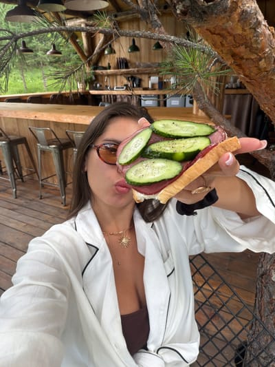 A young woman with dark hair, wearing sunglasses, a white collared shirt with black trim, and a brown top, holds a sandwich with cucumber and meat slices to her face, as if to take a bite. She is in an outdoor, rustic setting with wooden structures and furniture. The environment suggests a cafe or restaurant with a natural, wooded backdrop, possibly in the vicinity of Манский, Russia, indicated by the presence of pine trees and a wooden bar area. The image appears to be a selfie, with the woman looking directly at the camera.