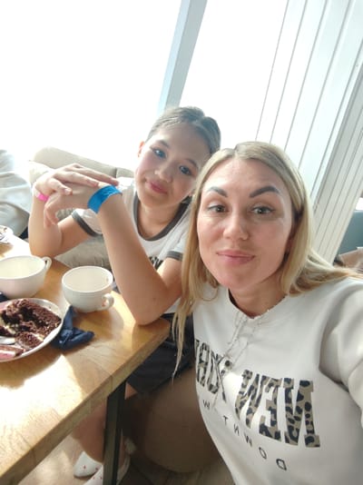 Two females, one adult and one child, are posing for a selfie at a table. The adult female has blonde hair and is wearing a white sweatshirt with a silver necklace. The child has dark hair and is wearing a white t-shirt with a black collar. They are both smiling and looking at the camera. On the table in front of them are two white cups, a plate of chocolate cake, and a dark blue napkin. The background is blurred, but appears to be an indoor setting with natural light. The location is Amursk, Russia.