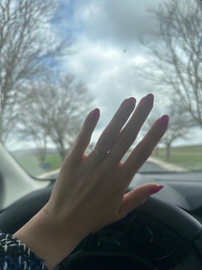 The image depicts a human left hand extended in the foreground, viewed from inside a vehicle in Izbul, Bulgaria. The hand exhibits manicured fingernails painted a magenta hue. A silver-toned ring with a single, clear, round-cut gemstone is present on the third digit (ring finger). A portion of a blue and white patterned woven garment is visible on the wrist. The upper section of a dark-colored steering wheel or dashboard, with partially visible illuminated instrument panel indicators, occupies the lower part of the frame. Through the vehicle's windshield, an outdoor environment is visible under a partly cloudy sky. The background features several leafless deciduous trees lining what appears to be a paved road or path that curves to the right. A flat green field extends to the left of the road. The scene suggests daylight during a cooler season such as late autumn, winter, or early spring.