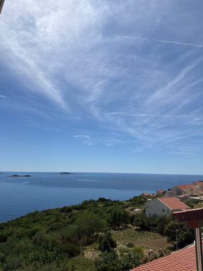 The image shows a view from a high vantage point in Soline, Croatia. The scene is divided into three distinct horizontal sections. The top section is the sky, which is a clear blue with wispy clouds and contrails. The middle section is the Adriatic Sea, a deep blue with slight ripples on the surface. Several small boats or islands are visible in the distance. The bottom section is the coastline, covered in lush green vegetation, interspersed with red-tiled rooftops of houses and a terraced garden. The scene suggests a bright, sunny day.