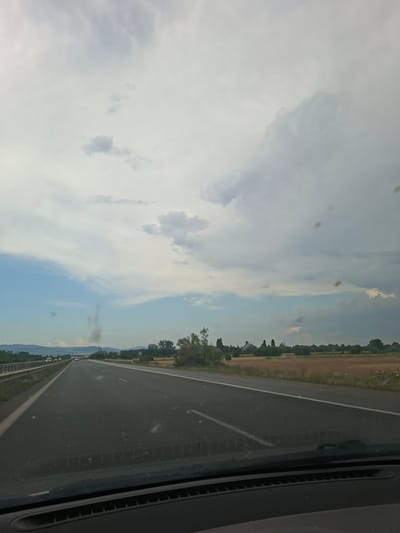 The image shows a view from inside a vehicle, looking out at a highway in Dolni Bogrov, Bulgaria. The perspective suggests the vehicle is in motion. The road stretches into the distance, bordered by a grassy shoulder and then a tree line. Beyond the trees, there are fields of dry vegetation and some distant hills or mountains. The sky is filled with clouds, ranging from light and wispy to darker, more substantial formations, suggesting a partly cloudy day. The time of day appears to be daylight, likely late morning or afternoon, given the brightness and shadows. There are a few small, dark specks on the windshield, which appear to be insects or dirt. Several vehicles are visible on the highway ahead, indicating moderate traffic. The overall scene conveys a sense of travel and a rural or semi-rural landscape. There is no visible text in the image.