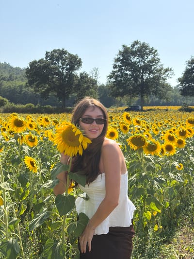 In a sunlit field of sunflowers, a young woman stands in the foreground, her head tilted slightly as she holds a single sunflower in front of her face, partially obscuring her features. She is wearing a white strapless top and a dark skirt. Her hair is long and brown, falling around her shoulders. She is also wearing sunglasses. The background is filled with a vast expanse of sunflowers stretching towards a treeline and hills. The sky is clear and blue. A dark-colored vehicle is visible in the distance, parked on what appears to be a dirt road. The setting is likely a rural area, given the expansive field and natural landscape. The image was captured in Montieri, Italy.