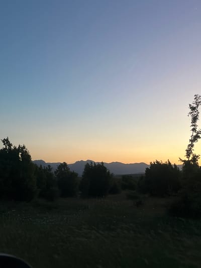 The image shows a landscape scene taken in Gorna Kremena, Bulgaria. The time appears to be around sunset, with a gradient sky shifting from blue to yellow. A mountain range is silhouetted against the sky. Trees are seen on the right side, partly obscuring the landscape. The ground is dark and grassy. No people or specific activities are visible.