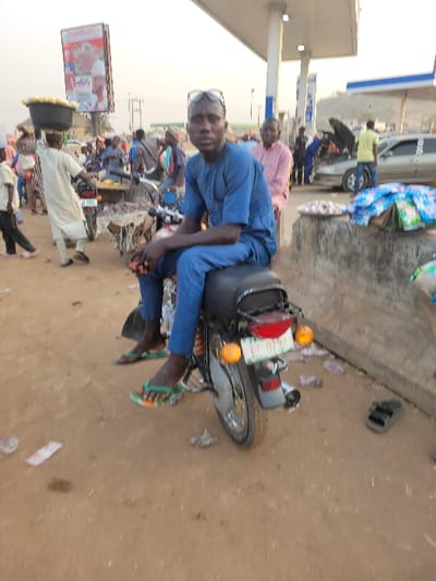 A man in blue attire, seated on a motorcycle, looks directly at the camera. The motorcycle is situated near a busy roadside scene with vendors, other people, and a gas station in the background. The road surface is predominantly brown, and litter is scattered nearby. The lighting appears to be daylight, creating shadows.