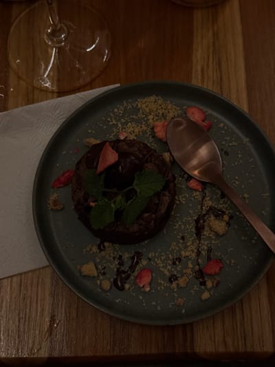 The image displays a dessert setting on a wooden table in a dimly lit indoor environment, likely a restaurant or bar in Guadalajara, Mexico. A circular, dark brown dessert, possibly a chocolate lava cake or brownie, sits centrally on a dark teal-colored ceramic plate. The dessert is topped with a scoop of dark ice cream or ganache, garnished with several fresh mint leaves and at least one piece of dried strawberry. The plate is scattered with light-colored crumbs and small pieces of dried strawberry, with drizzles of dark chocolate sauce. A copper-toned spoon rests on the right side of the plate. To the left of the plate, a folded white paper napkin is partially visible. In the upper left corner, the clear glass base of a wine glass is present. No individuals are visible. The specific location within Guadalajara is not discernible from the provided image.