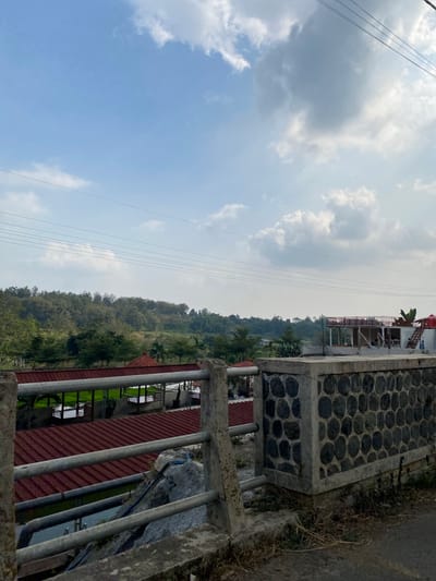 This is an outdoor, full-frame, vertical, eye-level shot of a rural landscape. The scene is dominated by a bright blue sky with scattered white clouds, and a lush green forested hill in the background. In the midground, there are several structures with red roofs, possibly farms or buildings. A stone-and-concrete wall with a metal railing in the foreground suggests this is a viewing point or a bridge. The overall impression is one of natural beauty and rural life in Kresek, Indonesia. There are no people visible in the frame.