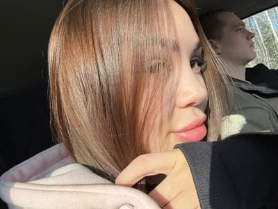 A young woman sits in a car wearing a pink hoodie with a black sleeve visible. She has light brown hair and full lips. Her gaze is directed to the right, away from the camera. In the background, a man is also seated in the car, his face in profile. Outside, trees are visible. The lighting is bright and sunny, casting shadows across the interior of the vehicle.