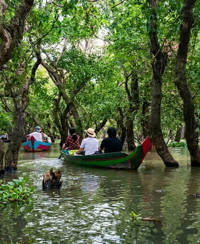 A group of people are enjoying a boat ride through a lush, flooded forest in the city of Soutr Nikom, Cambodia. Two boats are visible; the one in the foreground is green with red accents and carries three individuals. The person in the back is wearing a black shirt, the middle person is wearing a white shirt and a straw hat, and the person in the front is wearing a red and black checkered shirt. They are all facing away from the viewer, looking into the dense foliage. A second boat, painted with blue, pink, and yellow, is further ahead and to the left, carrying two people, also facing away. The water is calm and reflects the vibrant green of the trees that rise directly from it. Aquatic plants and a submerged piece of wood are visible in the water. The overall scene is one of serene exploration through a unique natural environment.