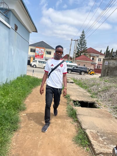 A young Black male walks toward the camera on a dirt path next to a concrete drainage ditch and overgrown grass. He is wearing a white t-shirt with red trim, black pants, black sneakers, and sunglasses. A black bag is slung across his chest. Behind him, a street is visible with cars and buildings, including a light blue building on the left with a sign that reads "MERCY SPRINGS INTERNATIONAL SCHOOL". The sky is cloudy. The location is identified as Abuja, Nigeria.