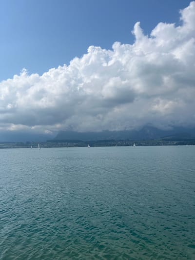 The image depicts Lake Thun in Switzerland under a sky transitioning from clear blue to heavy cloud cover. The foreground comprises the teal-blue surface of the lake, exhibiting small ripples and waves. Approximately seven small sailboats, characterized by white hulls and sails, are scattered across the lake from left to right, indicating movement. The mid-ground reveals the distant northern shore of Lake Thun, featuring a landscape of green hills, trees, and scattered buildings of a small town or village. The background is dominated by mountains, partially obscured by dense, dark gray cumulus and stratus clouds, particularly on the right side. The sky in the upper left quadrant shows patches of bright blue, with large, bright white cumulus clouds bordering the darker cloud mass. No individuals are discernible in the scene.