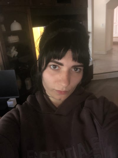 A single individual is depicted in an indoor residential setting in Santa Clarita, United States of America. The subject is an adult female with dark hair styled in an updo with bangs covering the forehead. She is wearing a dark brown hooded sweatshirt with the word "ESSENTIALS" embossed in a darker tone across the chest. Her face is directed towards the camera with a neutral expression. In the background to the left, a dark wooden shelving unit contains various objects, including a white dome-shaped item and stacked white rectangles. A dark computer monitor screen is partially visible in the lower left. To the right, a brightly lit hallway or room with light-colored walls and what appears to be tiled flooring is visible through an open doorway, leading to another interior space further back.