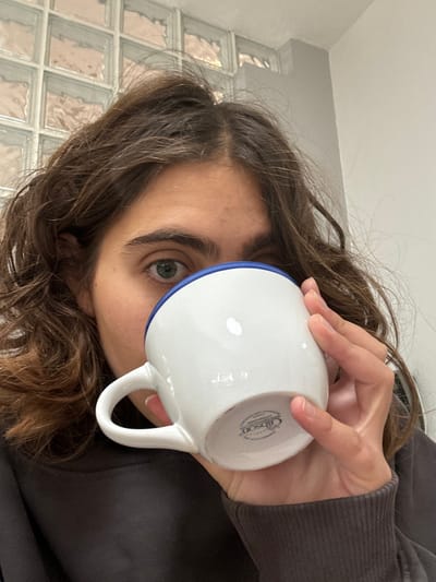 A close-up image depicts an individual partially obscured by a white ceramic mug with a blue rim, held to their mouth by their right hand. The person has dark, wavy hair, and their visible right eye is light-colored. They are wearing a dark long-sleeved garment. The mug's base bears a "Gibson" brand marking. The background consists of a wall section with translucent glass blocks on the upper left and a plain, light-colored wall on the upper right. No visual identifiers are present in the image to determine its location within the city of Menashe Regional Council, Israel.