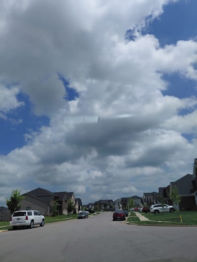 The image depicts a residential street in Lexington, Kentucky, USA, on a cloudy day. The street is lined with modern two-story houses, mostly brick and siding, with dark-colored roofs. Several cars are parked on the street and driveways, including a white SUV, a red sedan, and a white sedan. The sky is filled with a mixture of white and gray clouds, with patches of blue sky visible. Green lawns and trees are present in front of the houses. The perspective is from a car driving down the street.