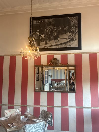 This media file captures an interior scene, likely within a restaurant or cafe in Simon's Town, South Africa. The image is taken from a slightly high and rotated perspective, showcasing various decorative elements and a set table.

On the left, a large, black-framed black and white photograph is prominently displayed on a light-colored wall. The photograph depicts a street scene with multiple people, including one individual on what appears to be a scooter or moped. Visible text on the photograph identifies it as "Ruth Orkin" and "American Girl in Italy 1951." Below the photograph, an ornate chandelier with clear, crystal-like embellishments and multiple illuminated bulbs hangs, casting a warm glow.

To the right, a wall is adorned with striking horizontal stripes of alternating red and white. An ornate, antique-style gold-framed mirror is mounted on this striped wall. The mirror's reflection clearly shows a person holding a smartphone, taking a selfie of themselves and the interior. The reflection also reveals more of the inner space, including a wall with blue and white stripes further inside.

In the lower right portion of the image, a rustic wooden table is meticulously set for two. Each setting includes a wine glass, cutlery, and a decorative cloth napkin or placemat; one features a floral pattern, while the other displays a gray and white damask design. A small, white sign with the word "Reserved" is visible on the table, indicating its unavailability. The overall setting suggests a charming and thoughtfully decorated dining establishment.