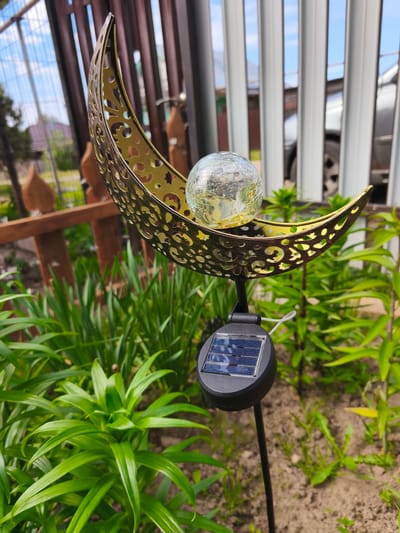 A close-up, eye-level shot features a solar-powered garden light in the shape of a crescent moon with a crackled glass ball at its center. The moon is a metallic gold color and has intricate cut-out patterns. Beneath the moon, a black solar panel on a stake is visible. The garden light is positioned amongst lush green plants with pointed leaves. In the background, a brown wooden fence separates the garden from a light-colored, vertical-slatted fence behind which a car is partially visible. The sky above is blue with scattered white clouds. The scene appears to be a residential garden in a city, possibly Puchezh, Russia, given the overall aesthetic and the mention of Russian location. There are no people or animals present in the image. The lighting suggests it is daytime, with sunlight illuminating the scene.