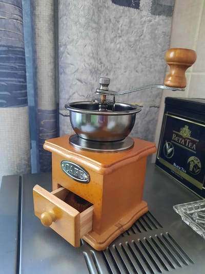 The image displays a vintage-style wooden coffee grinder with a metal hopper and a wooden handle, positioned on a stainless steel surface. A drawer is partially open at the base of the grinder. To the right of the coffee grinder, a black tin of "Beta Tea" is visible. The background consists of vertical curtains with a blue and gray pattern. The scene is illuminated by ambient light, creating soft shadows. There are no people visible in the image. The coffee grinder and tea tin suggest a domestic or kitchen environment, possibly in Зеленогорский, Russia, given the prompt's context.