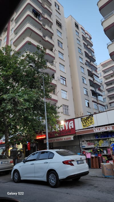 The image shows a street scene in Çukurova, Turkey, with a modern apartment building as the central focus. The building is tall and cream-colored with multiple balconies, many of which have dark red trim and metal railings. Clothes are hanging on a clothesline on one of the upper balconies. A large, leafy green tree is situated in the foreground on the left, partially obscuring the lower floors of the building. A street lamp is visible next to the tree. Below the apartments, a row of storefronts lines the street. One prominent storefront has a red and white sign that reads "KAYA EV TEKSTİLİ & ÇEYİZ." To the right of this, a yellow sign with black text says "SATILIK" (for sale) and a phone number. Further to the right, another storefront displays various products, including buckets and containers, with a sign that appears to read "YALIMLAR." A white sedan is parked on the street in front of the storefronts. The sky above is a clear, pale blue, suggesting it is either early morning or late afternoon.