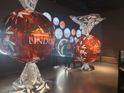 The image was taken in Kilchberg (ZH), Switzerland. The scene features two oversized Lindor chocolate installations in a Lindt chocolate museum. These are large, spherical representations of the iconic Lindor chocolate wrapped in red foil and clear cellophane-like material at the top and bottom. One installation has a white floral pattern on the clear cellophane, while the other has a clear, white-patterned extension resembling the foil wrapping. In the background, there is a wall adorned with circular images, possibly of people. Two individuals stand between the two chocolate installations. The person closer to the right side wears a Red Bull Racing jacket. The floor is glossy. The overall environment appears to be a museum or exhibit space.