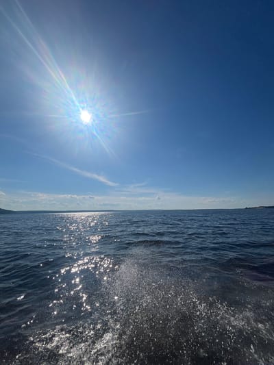 The image depicts a bright, sunny day over a body of water, likely a lake or sea, given the distant landmass visible on the horizon. The sun is intensely bright, casting a prominent lens flare with a rainbow effect across the upper left quadrant of the sky, which is a clear, deep blue. The water is dark blue with whitecaps and sparkling reflections of the sun. In the foreground, on the right side, a splash of water is visible, suggesting movement, perhaps from a boat or wave action. The environment is natural and serene, with a clear sky and calm water. The location is identified as Siukterka, Russia.