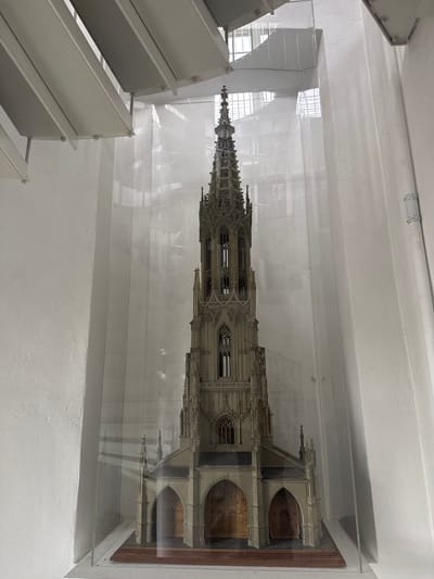 A vertical, eye-level, medium shot captures a detailed architectural model of a cathedral spire displayed inside a clear acrylic case. The model is intricately designed with Gothic architectural elements, including pointed arches, ornate carvings, and numerous spires. It appears to be made of a light-colored material, possibly plaster or resin, and is illuminated from above, casting subtle shadows. The model is positioned horizontally within the case, extending from the left to the right of the frame.
The setting is indoors, with white, textured walls and a white ceiling visible in the background and foreground. A section of a modern, angular white staircase or tiered structure is present in the lower left quadrant of the image, suggesting the model is on display in a museum, exhibition space, or public building. The acrylic case reflects light, creating a hazy effect and obscuring some of the finer details of the model and the background. The overall impression is one of a well-preserved or newly constructed model, highlighting the craftsmanship and grandeur of Gothic architecture. No people are visible, and the scene suggests a static display rather than an active event. The time of day and weather are not discernible from the image. There is no visible text. The location context is Bern, Switzerland.