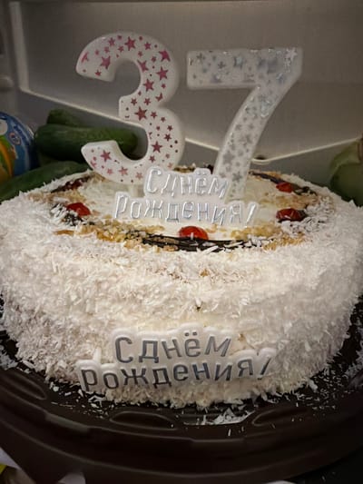 A white cake decorated with shredded coconut and red berries sits on a dark plate. Two candles shaped like the numbers "3" and "7" are placed on top of the cake. The "3" candle is white with pink stars, and the "7" candle is white with silver glittery stars. The cake also has a white inscription that reads "С Днём Рождения!" (Happy Birthday!) in two lines. In the background, partially visible, are cucumbers and what appears to be a blue and yellow toy. The image was taken in the city of Subkhankulovo, Russia.
