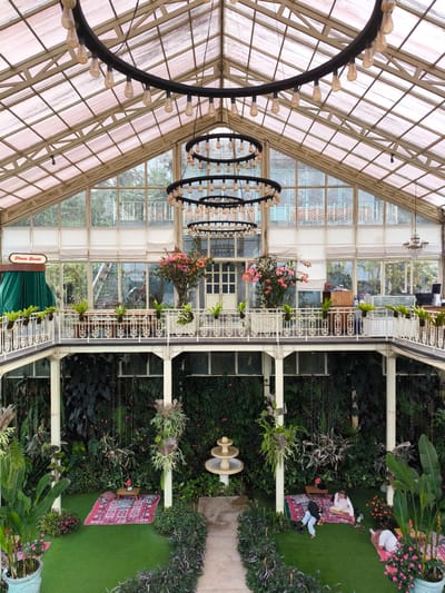 Captured from an elevated vantage point inside a spacious greenhouse-like building, the scene reveals a multi-tiered, lush interior. The structure boasts a glass-paneled roof and walls, allowing ample natural light to permeate the space. Massive circular chandeliers with numerous bulbs hang from the ceiling. A second-story balcony lined with plants overlooks the ground floor, which features artificial turf and a variety of plants and ferns. On the ground level, there are red rugs laid out with small tables, suggesting a lounge area. People are sitting and reclining on the rugs, engaging in leisurely activities. A stone pathway leads to a small fountain in the center of the ground floor. The "Photo Booth" sign is visible on the upper balcony. The location is in Candikuning, Indonesia.