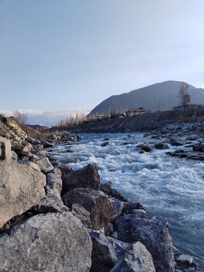 This image depicts a wide, rocky river flowing through a mountainous valley in Qoraqiya, Uzbekistan. The foreground is dominated by large, irregular gray boulders and smaller stones covering the left bank. The river occupies the central and right portions of the frame, characterized by turbulent, white-capped water indicative of a swift current, with numerous partially submerged rocks creating ripples and foam. Along the elevated right bank, a line of bare deciduous trees runs parallel to the river. Behind these trees, several residential-type structures are visible, including a prominent building with a reddish-brown roof. A large, dark-colored mountain forms the backdrop on the right, rising significantly above the river and structures. The sky overhead is clear and light blue, transitioning to a slightly darker shade higher up. In the far background, to the left, distant mountain peaks or low clouds are faintly visible. No individuals or specific activities are depicted.