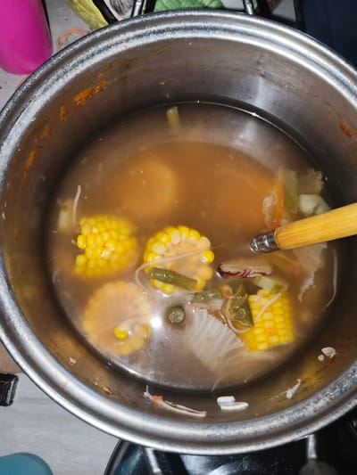 A top-down, eye-level, medium shot captures a stainless steel pot filled with vegetable soup. The soup contains pieces of corn on the cob, green beans, and cabbage. A wooden spoon with a metal tip rests in the soup, submerged in the broth. The pot's interior shows some staining and residue, particularly around the rim. The background is blurred and out of focus, but hints of a countertop and possibly other household items are visible. There are no people present in the image. The scene appears to be indoors, likely in a kitchen environment. There is no information to suggest a specific location in Barengkrajan, Indonesia.