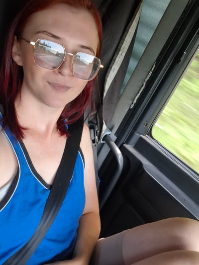A medium shot captures a woman with red hair and square-framed glasses, wearing a blue tank top and a seatbelt, inside a vehicle. The woman is looking towards the camera, with her mouth slightly open and a neutral expression. Reflections of a road and trees are visible in her glasses. The interior of the vehicle shows dark gray panels and a blurred view of greenery through the window, indicating movement. The scene appears to be set on a road, possibly in or near Sukhoy Ruchey, Russia, given the natural landscape visible outside.