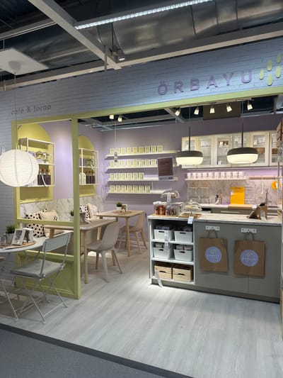 The image depicts a cafe or food stall area within a commercial establishment, possibly a department store or supermarket, given the retail display setup. The scene is brightly lit and features a modern, clean aesthetic with a color palette of light purple, yellow, and light wood tones.

In the foreground, there is a counter area with shelves holding white storage bins and two brown paper shopping bags hanging on the front. The bags are decorated with a circular logo featuring droplets and the text "ORBAYU cafe & forno". Above the counter, a kitchen or display area is visible, with cabinets, glassware, and various kitchenware.

To the left, a seating area is set up with a bench upholstered in light fabric and decorated with patterned cushions. Two small tables are present, one with white folding chairs and another with light wood chairs. Shelving units hold decorative items and what appear to be coffee-making equipment and bottled goods.

On the wall behind the seating area, a series of yellow square frames with text are displayed, likely menu items or decorative captions, including "cappucce" and "expres". Above this, a large sign with purple lettering spells out "ÖRBAYU" with decorative yellow droplet shapes.

The floor is a light-colored wood-effect laminate. The overall environment suggests a welcoming and stylish cafe or food service area designed for customer engagement and product display. The location is identified as Siero, Spain.