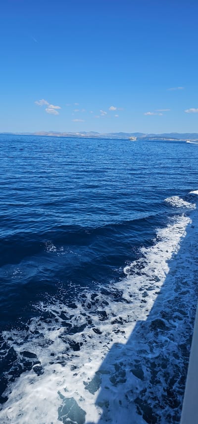 This is a medium shot, horizontal-format image taken from a boat moving through the sea. The water is a deep, rich blue, with visible waves and white foam generated by the boat's passage. A shadow from the boat's structure falls across the water on the right side of the frame, highlighting the texture of the churning sea. In the distance, a coastline is visible, lined with mountains. A white ferry or larger boat is also visible on the horizon. The sky is a clear, bright blue with a few small, scattered clouds. A faint contrail from an airplane is visible in the upper left portion of the sky. The overall impression is of a bright, sunny day at sea, with the camera looking towards the distant land. The scene evokes a sense of travel and a peaceful day outdoors. There is no visible text.