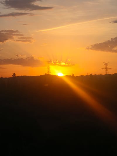 The image captures a sunset over a rural landscape, likely on the outskirts of Riba-roja de Túria, Spain. The sky is a vibrant orange and yellow, with rays of sunlight emanating from behind the horizon. Clouds are scattered across the sky, some tinged with pink and orange. Silhouetted against the bright sky are several power line towers, indicating the presence of infrastructure. The foreground is a dark silhouette of rolling hills and trees, suggesting a natural environment. The overall impression is one of natural beauty and tranquility.